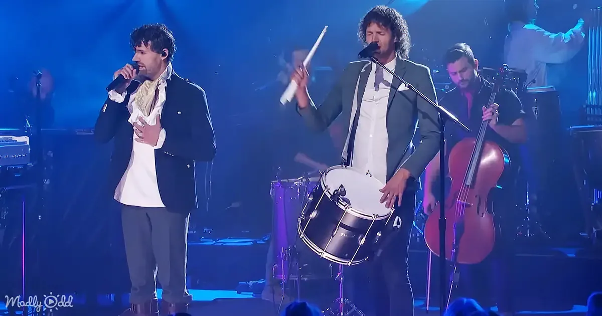 Two men sing on stage, one with a microphone, the other with a drumstick, accompanied by a band.