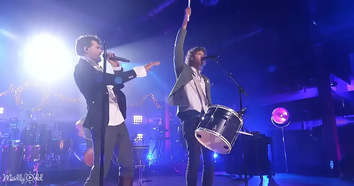 Two musicians perform on stage, one singing into a microphone, the other playing a drum, under vibrant stage lighting.