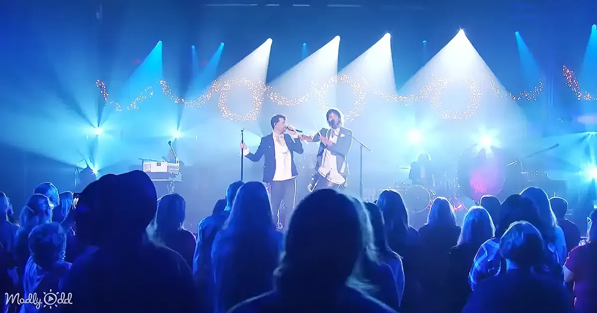 Two performers sing on stage with bright lights and a festive backdrop, surrounded by an audience.