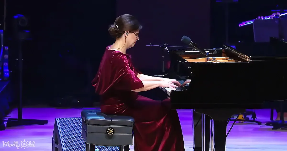 A woman in a red dress plays a grand piano on stage under purple lighting.