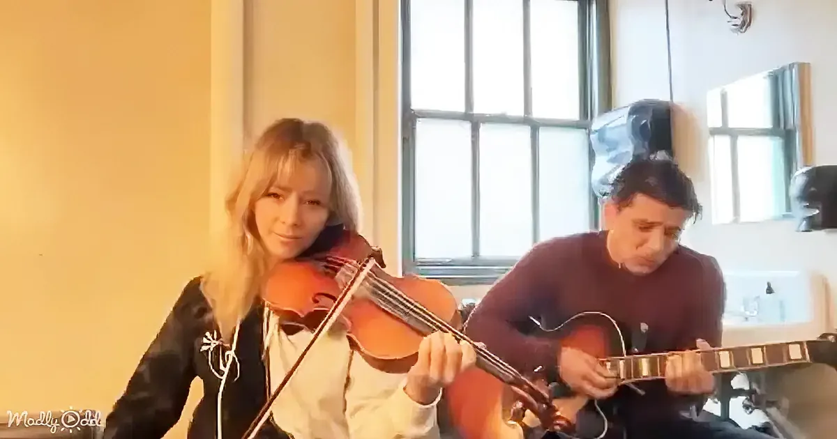 A woman plays violin while a man plays guitar in a warmly lit room.