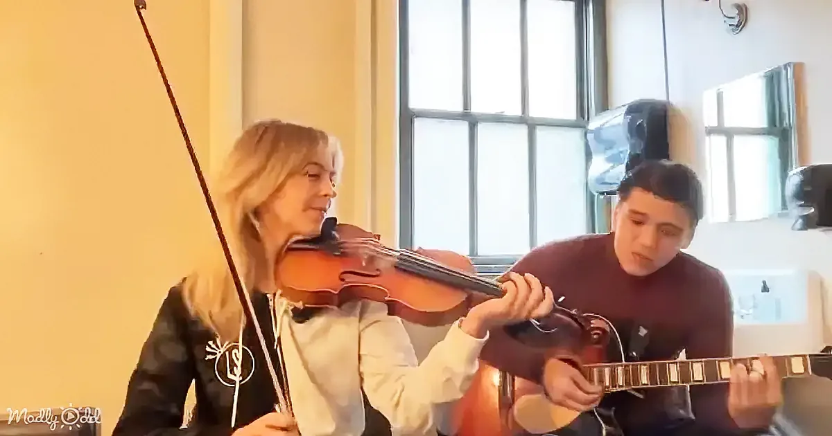 A woman plays violin while a man plays guitar in a room with large windows.