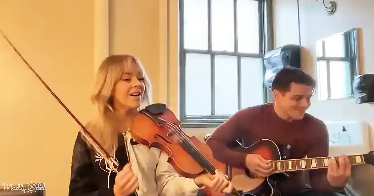 A woman plays violin while a man plays guitar, both smiling, in a warmly lit room.
