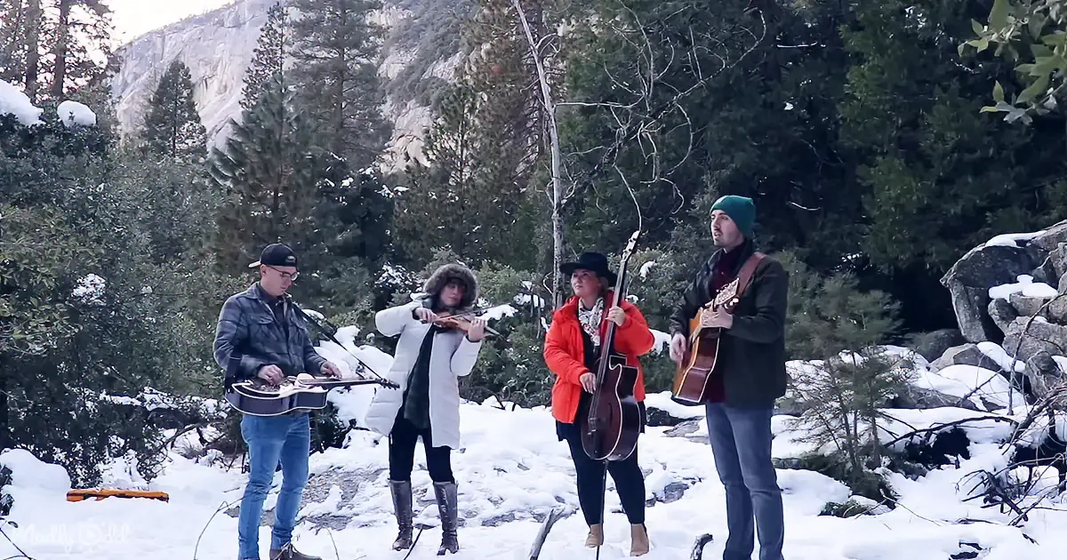 A group of musicians plays in a snowy forest, surrounded by snow-covered trees and mountains.