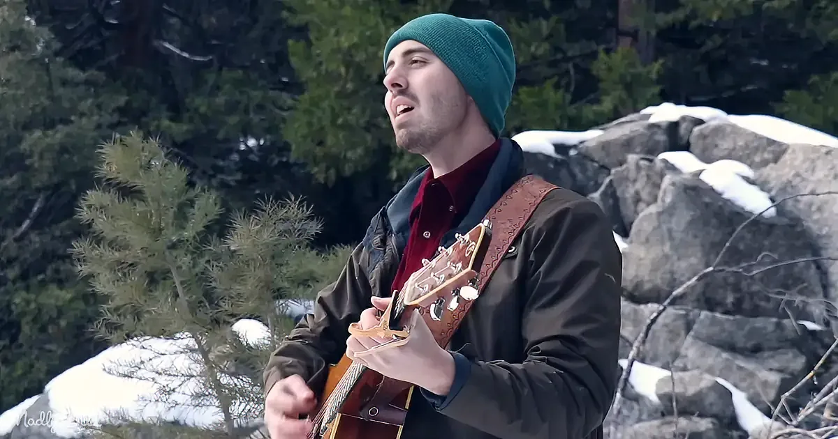 A man in a green beanie and brown jacket plays a guitar outdoors, surrounded by snow and trees.