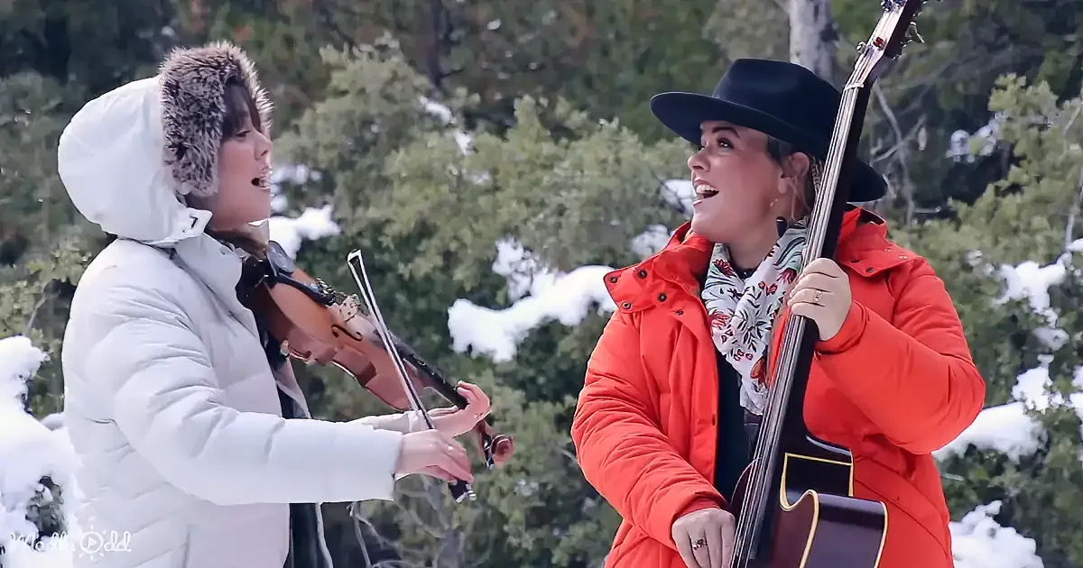 Two women in winter attire play violin and guitar in snowy forest.