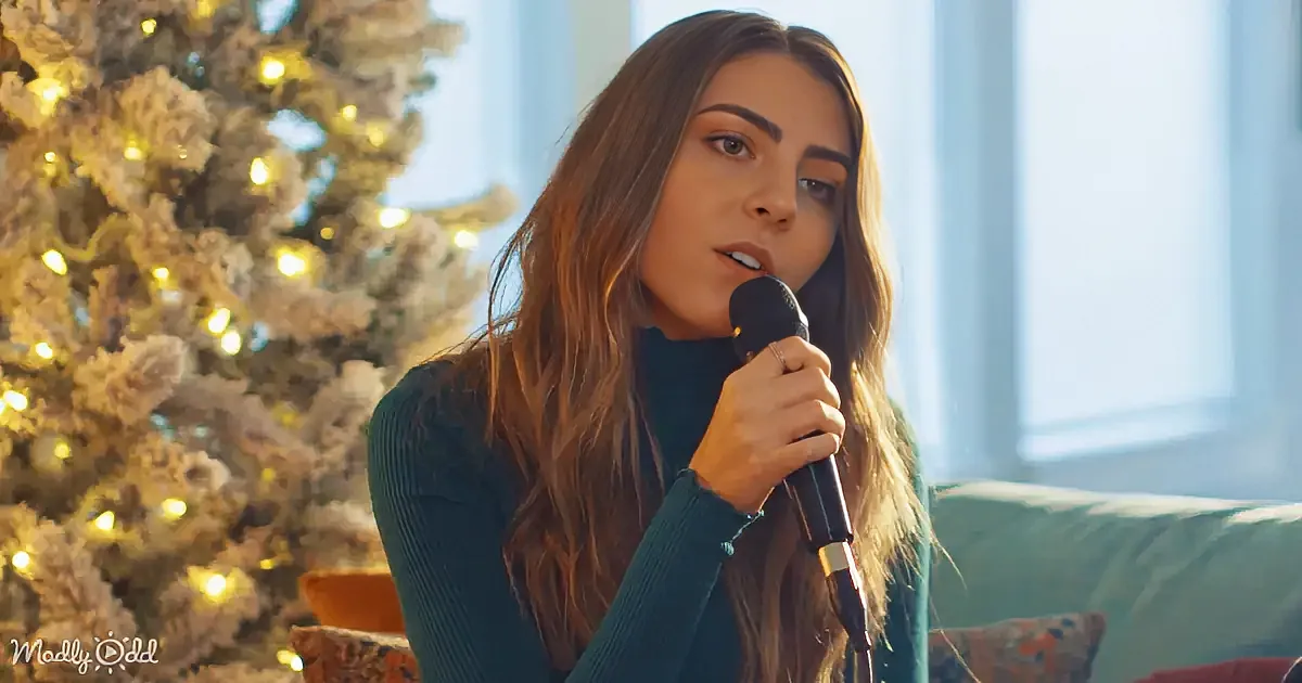 A woman with long hair holds a microphone, standing in front of a decorated Christmas tree.