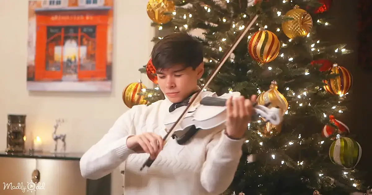 A boy in a white sweater plays the violin in front of a decorated Christmas tree.