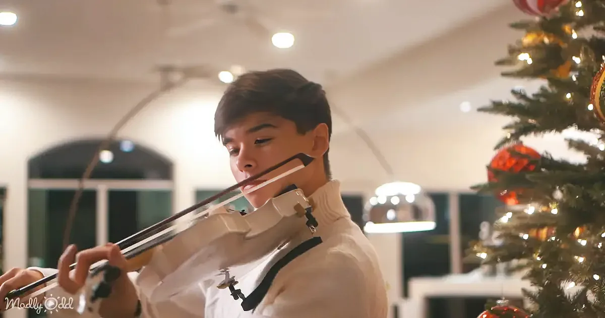 A person in a white shirt plays the violin near a decorated Christmas tree.