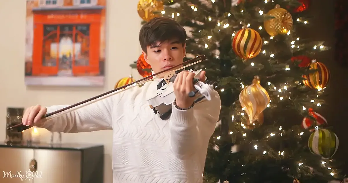 A person in a white sweater plays the violin in front of a decorated Christmas tree.