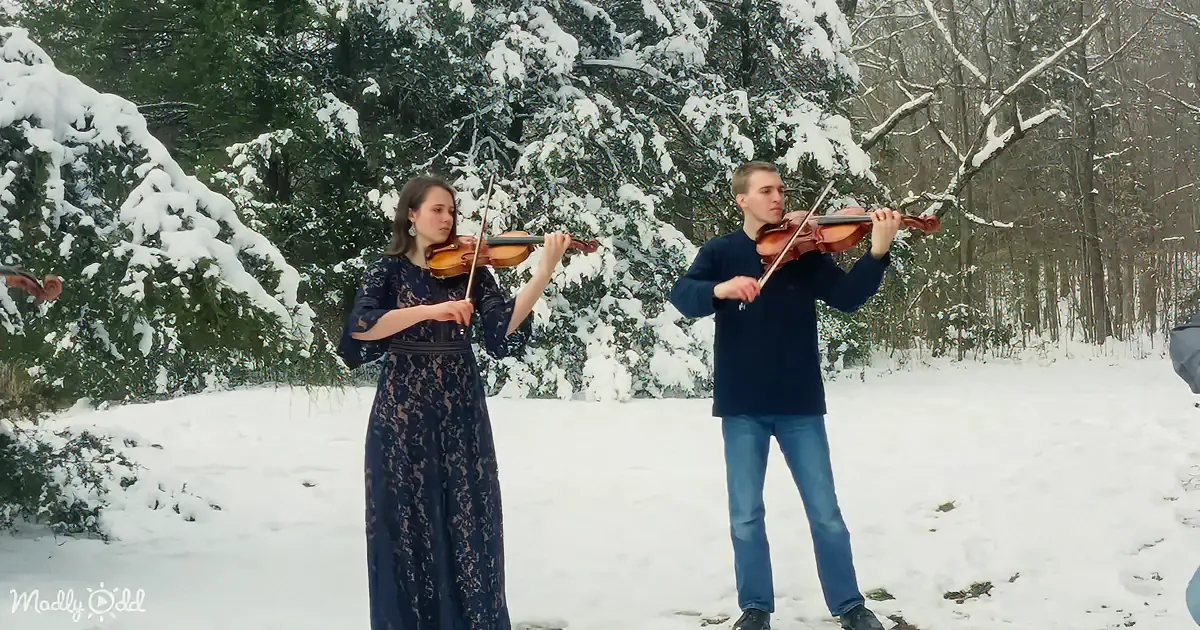 A woman in a lace dress and a man in jeans play violins in a snowy forest.