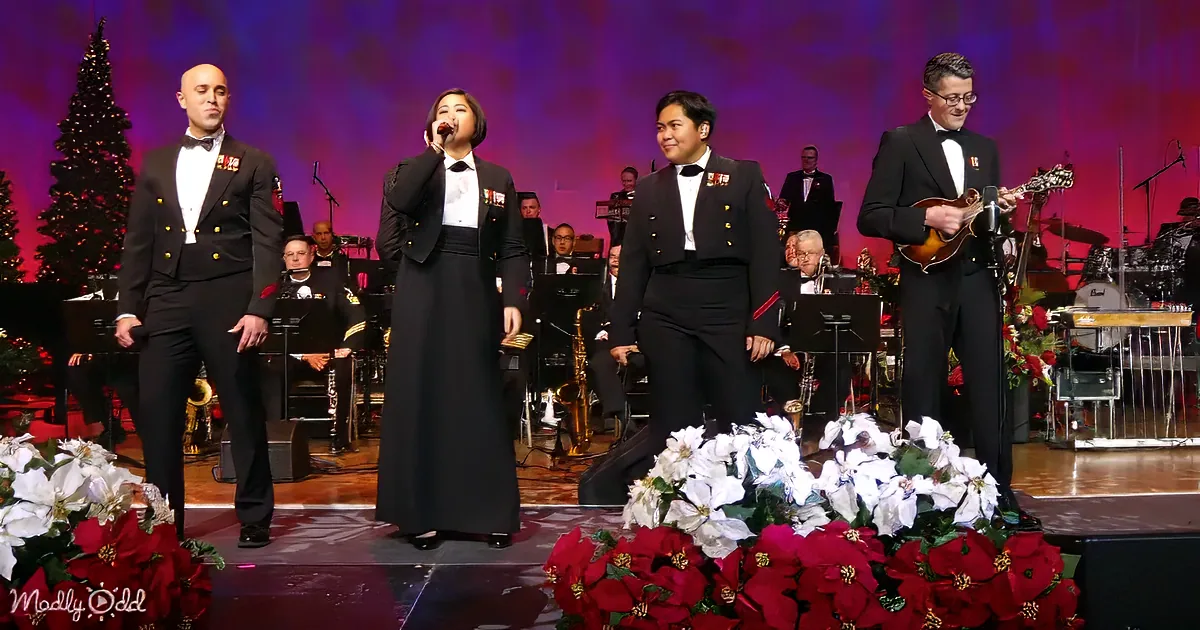 A group of performers in formal attire sing on stage with a Christmas tree and flowers in the foreground.