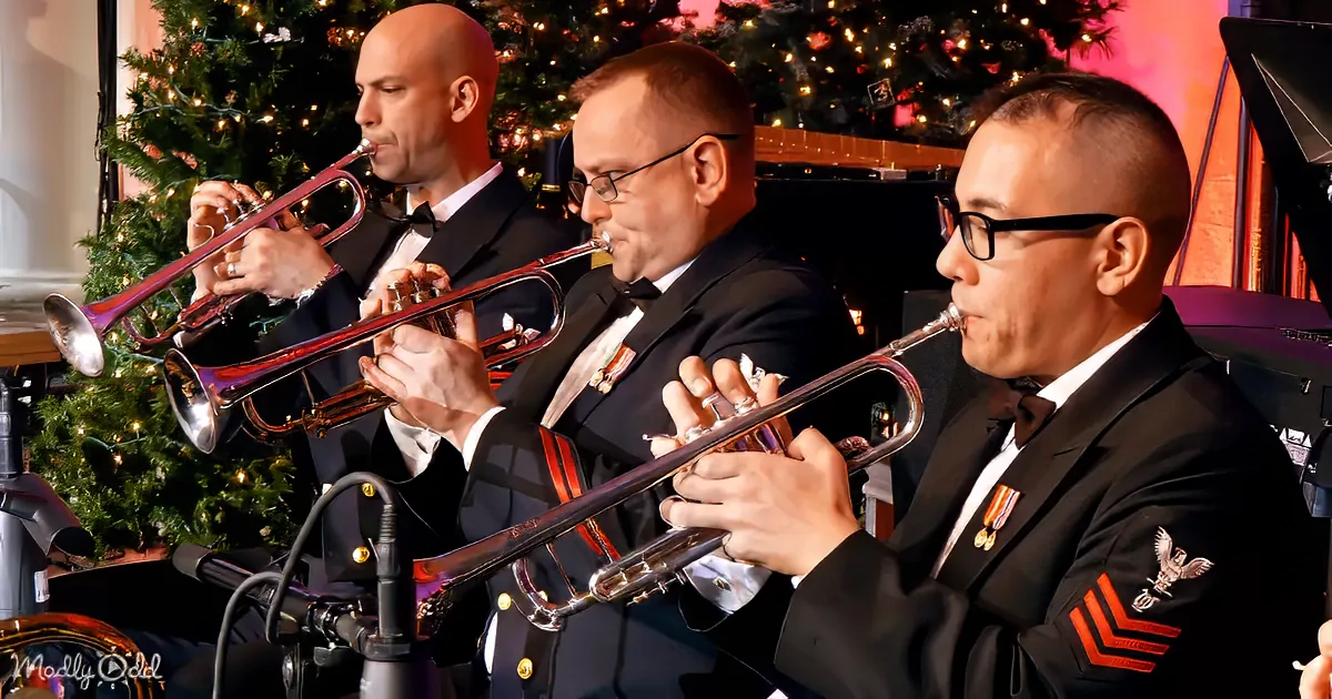 Three men in formal attire play trumpets in front of a decorated Christmas tree.