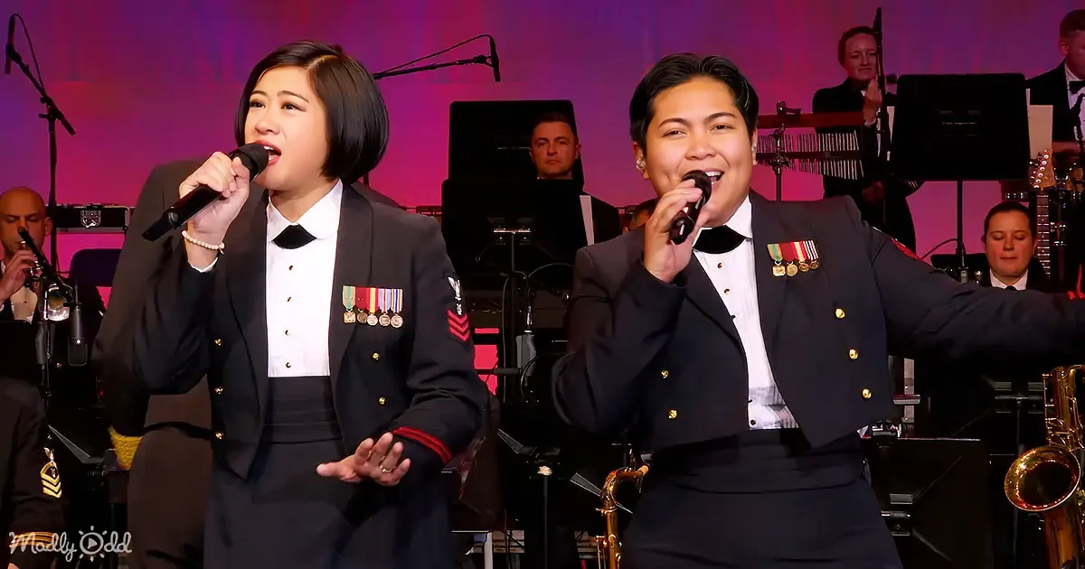 Two women in military-style uniforms sing into microphones, surrounded by musicians and instruments.