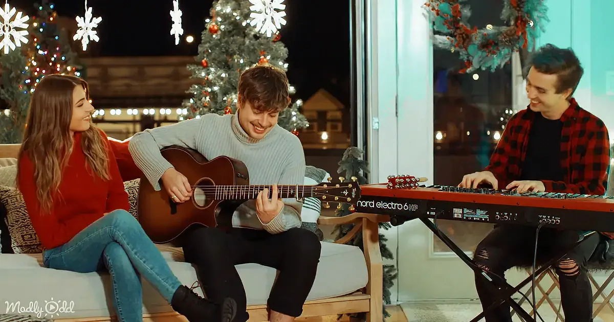 A man plays guitar while a woman and another man smile, surrounded by Christmas decorations.