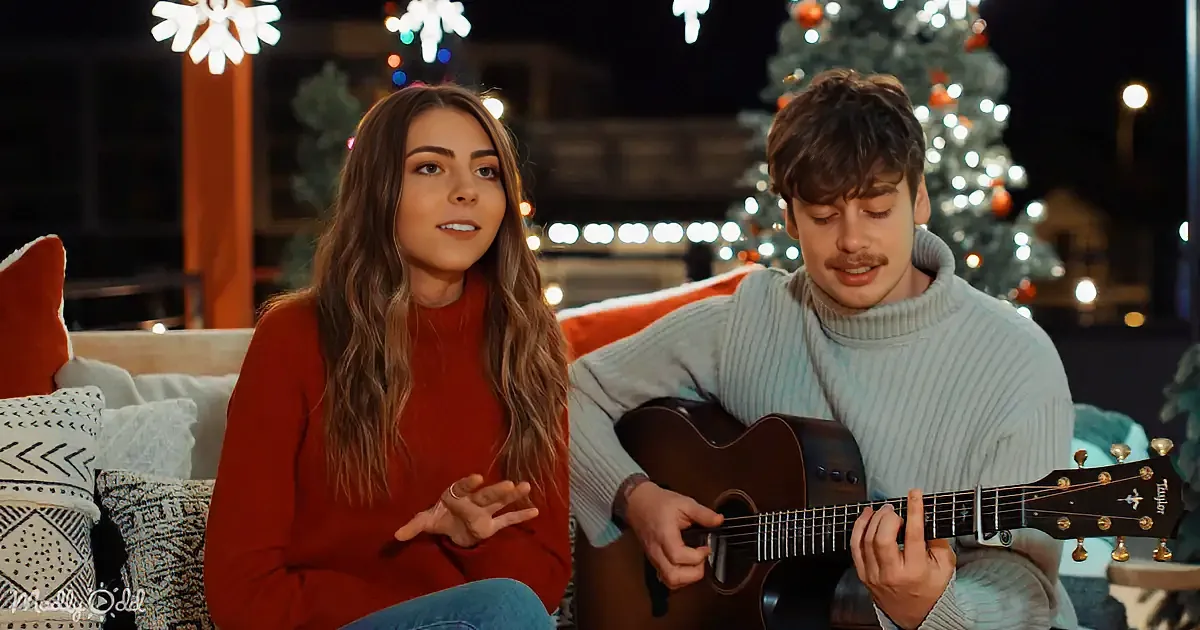 A woman in red and a man in a sweater play guitar and sing in front of a Christmas tree.