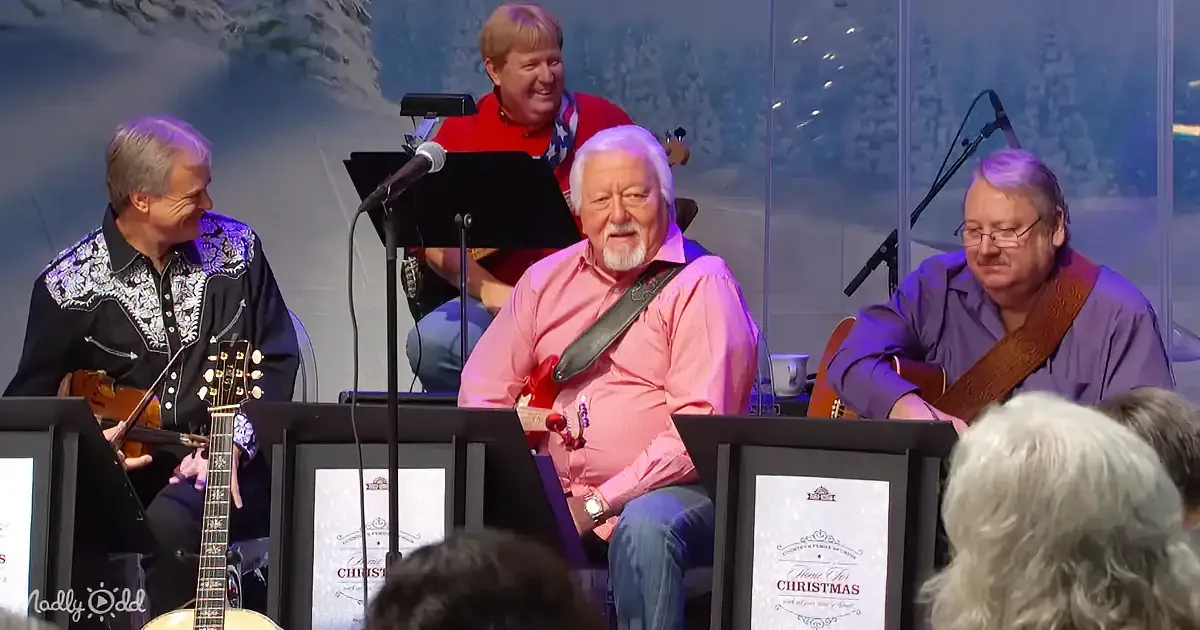 A man in a pink shirt plays guitar on stage with other musicians, smiling audience in front.