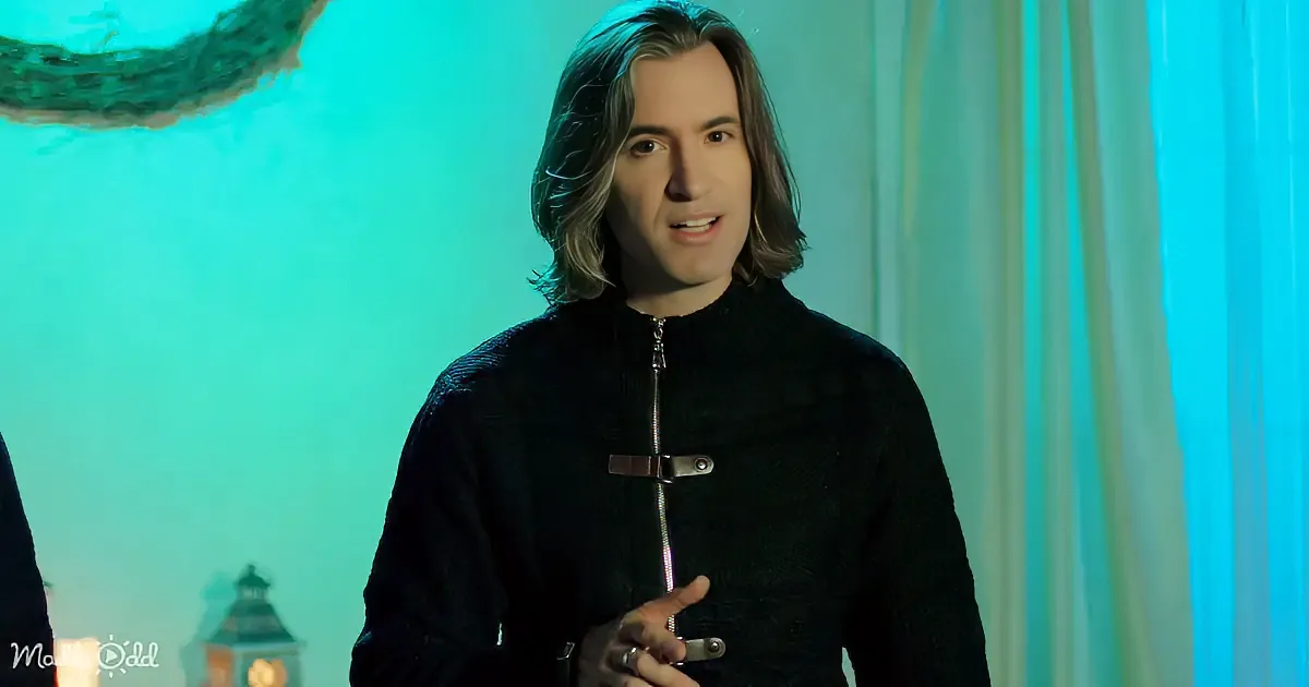 A person with long hair, wearing a black zip-up sweater, stands in a room with teal lighting and a lantern in the background.