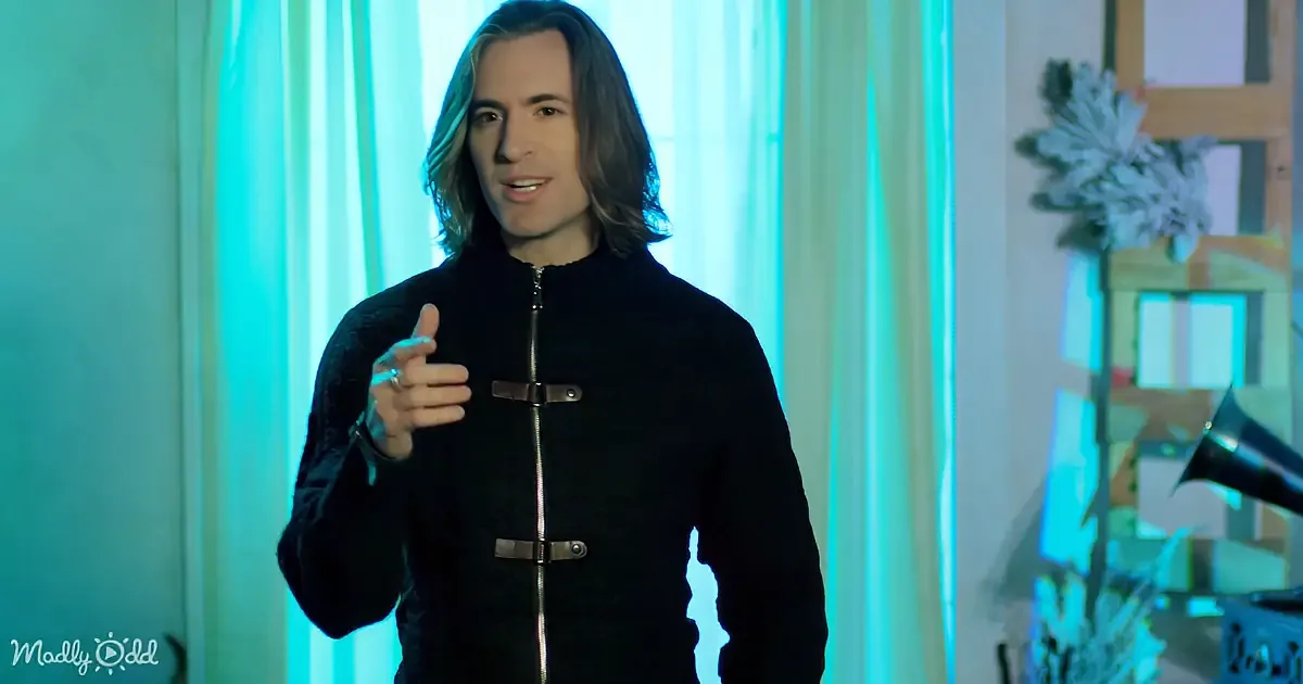 A person with long hair, wearing a black jacket, gestures with their right hand in front of a blue-lit background.