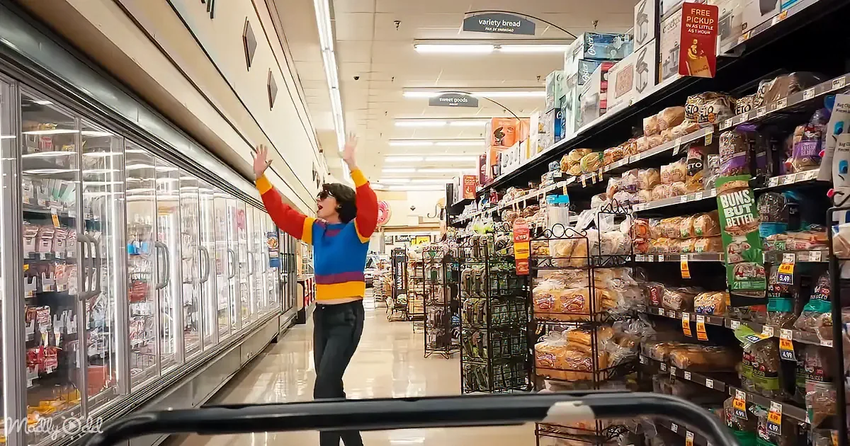 A person in a colorful sweater stands in a grocery store aisle, arms raised, with a refrigerator and bread shelves visible.