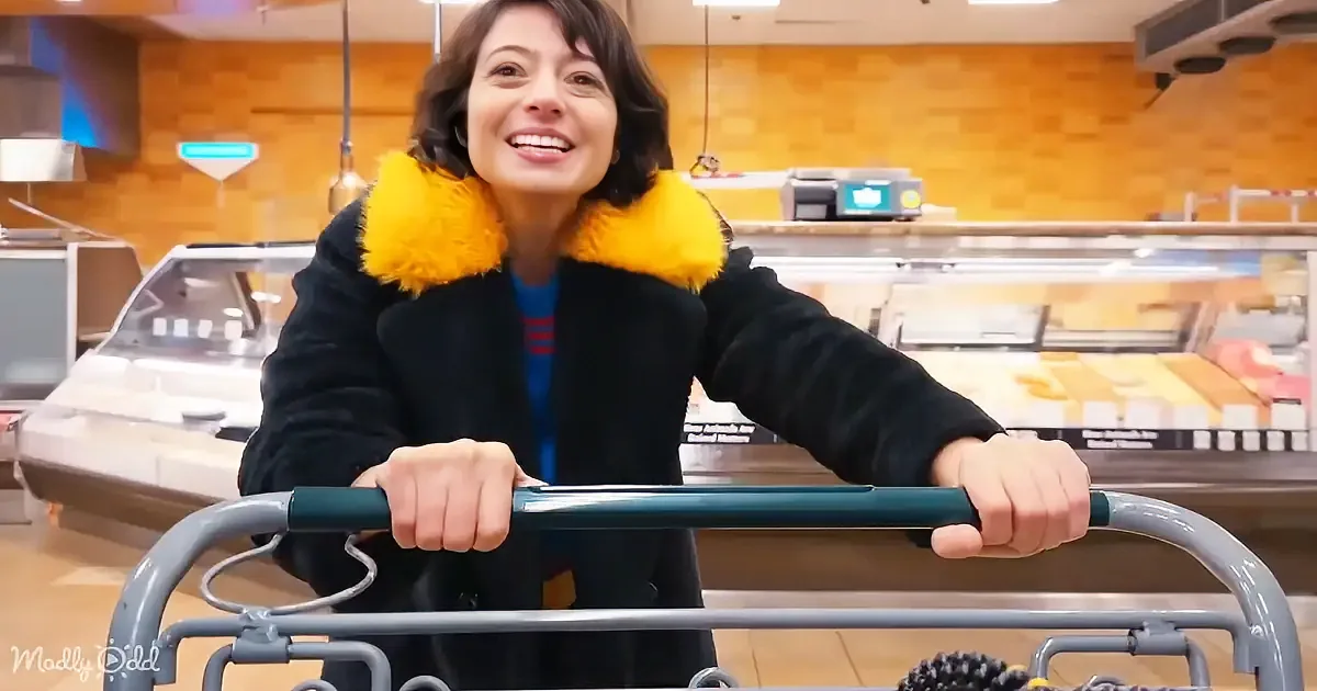 A woman in a black coat with a yellow fur collar smiles while pushing a shopping cart in a grocery store.