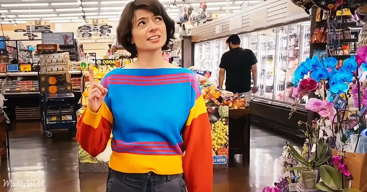 A woman in a colorful sweater points upwards in a grocery store aisle with produce and flowers.