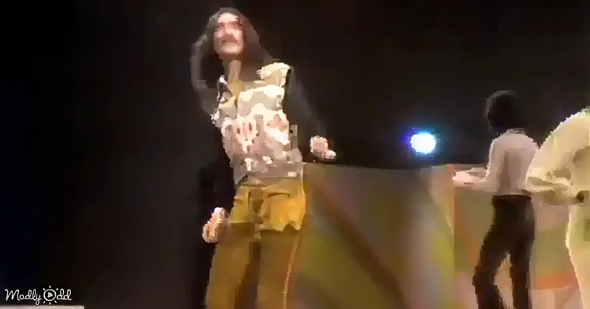 A person in a camouflage shirt and yellow pants stands on stage, holding a microphone, with a bright light in the background.