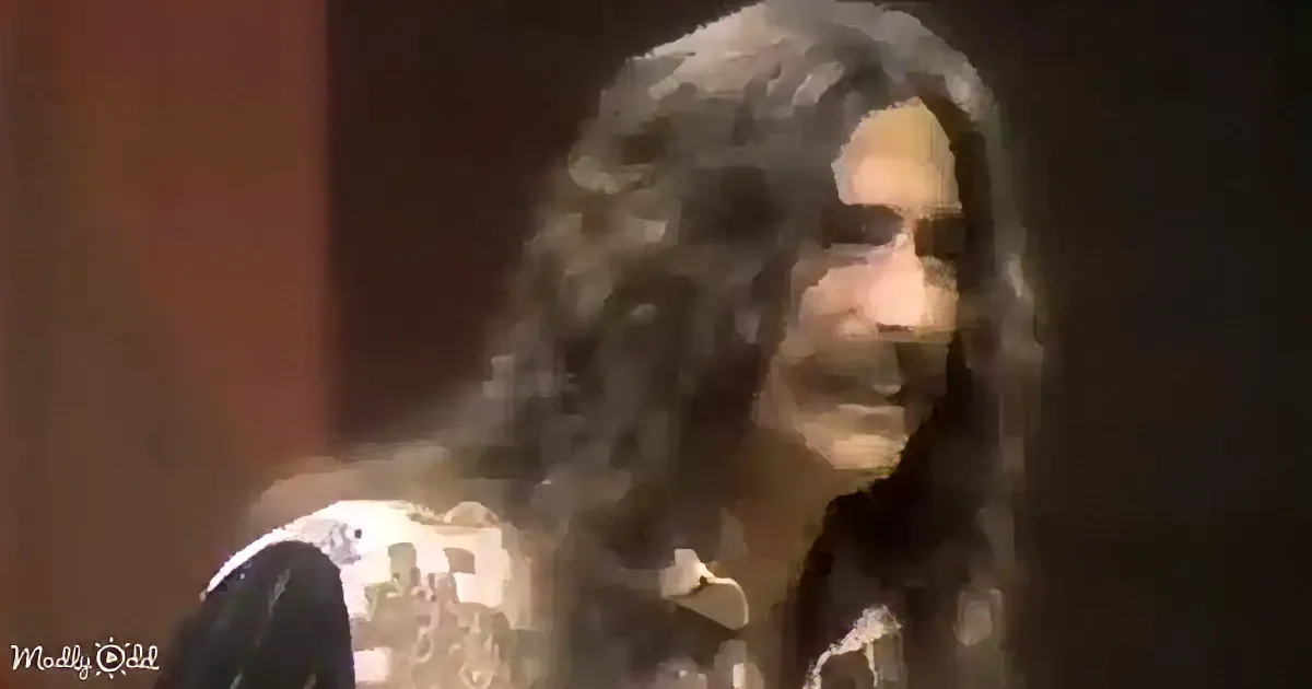 A person with long, curly hair and a patterned shirt is seen in a dimly lit setting.