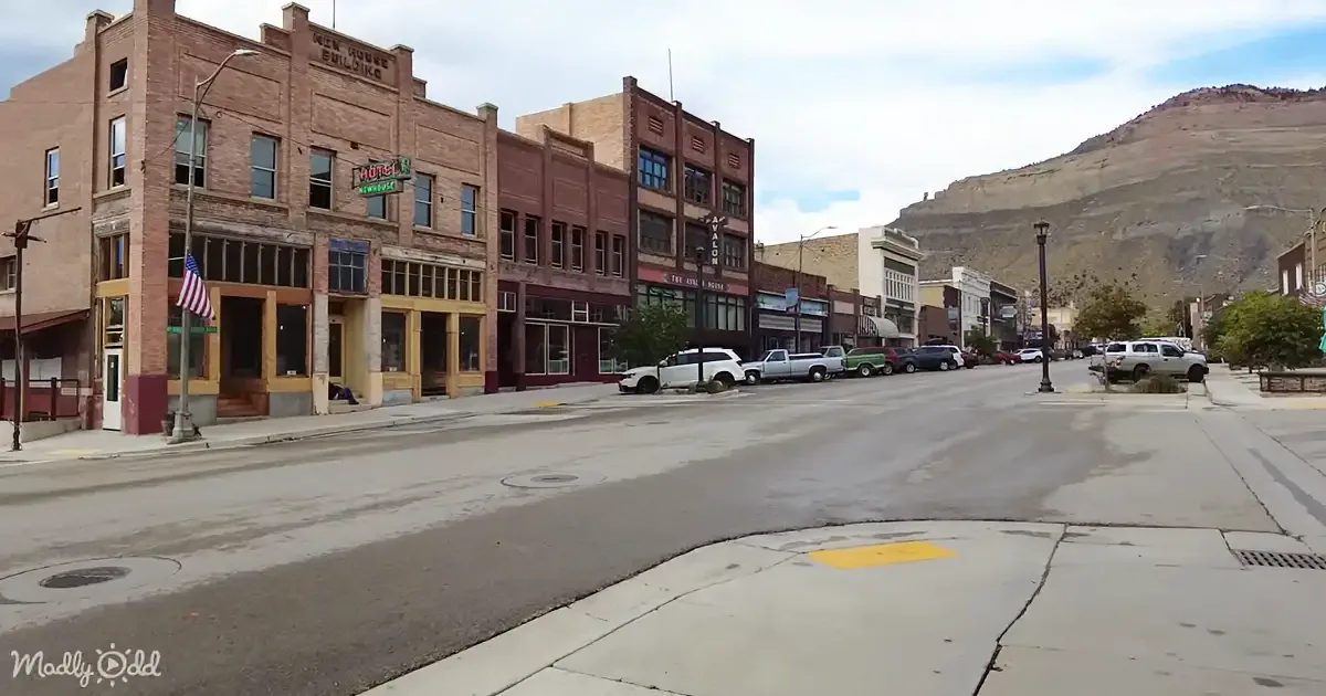A quiet town street with old brick buildings, parked cars, and a mountain in the background.