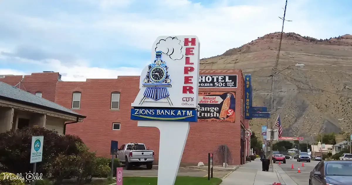 A sign for "Helper" with a train design stands in front of a brick building with a hotel sign and a mountain in the...