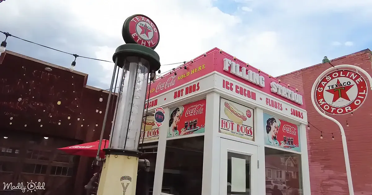 A vintage Texaco gas pump stands in front of a pink filling station selling Coca-Cola, hot dogs, and ice cream.