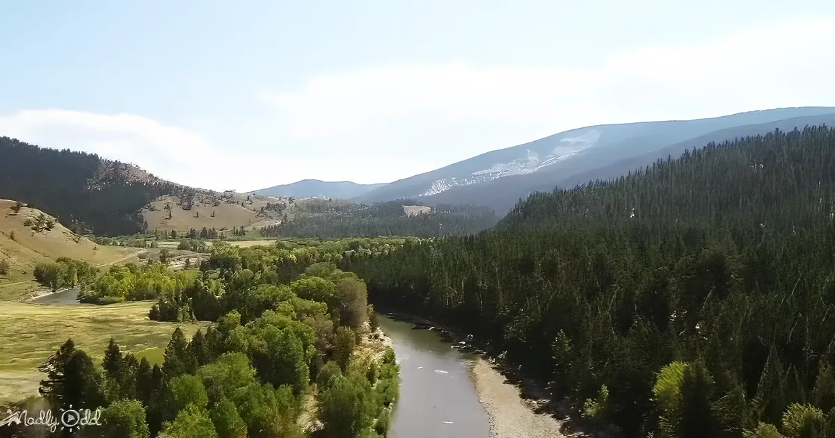 A winding river flows through a lush forested valley, surrounded by rolling hills and distant mountains under a partly...