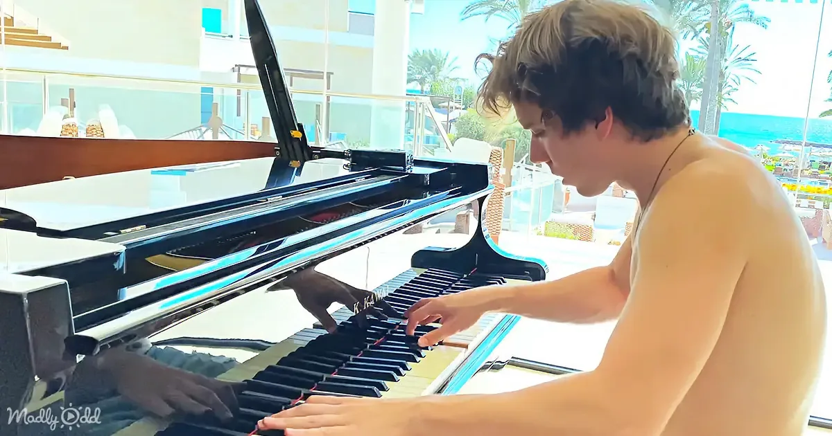 A shirtless man plays a grand piano by a beachside, with palm trees and ocean in the background.