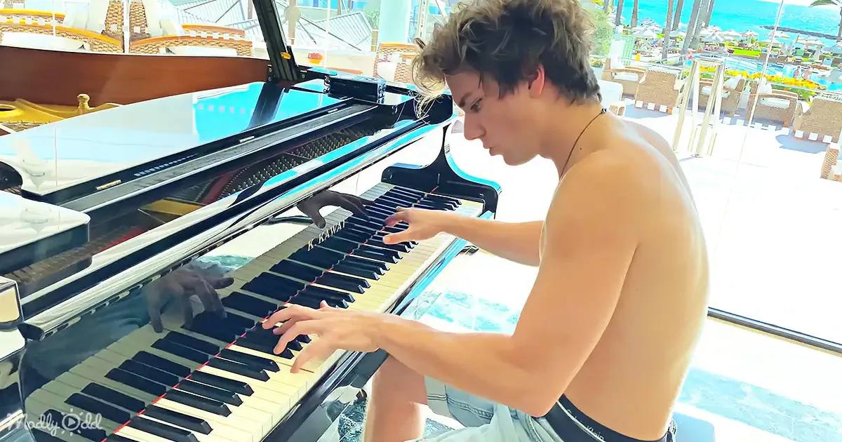 A shirtless man plays a grand piano on a sunny day by the beach.