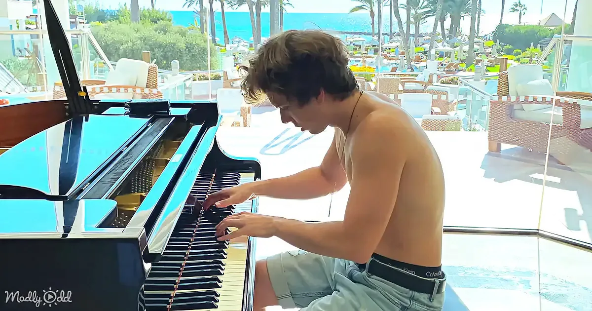 A shirtless man plays a grand piano on a sunny terrace overlooking a beach.
