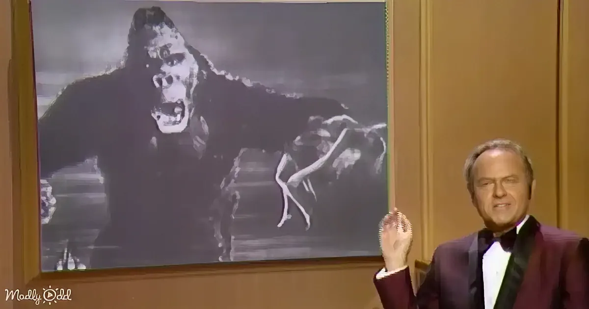 A man in a tuxedo gestures while a large gorilla image is projected behind him.