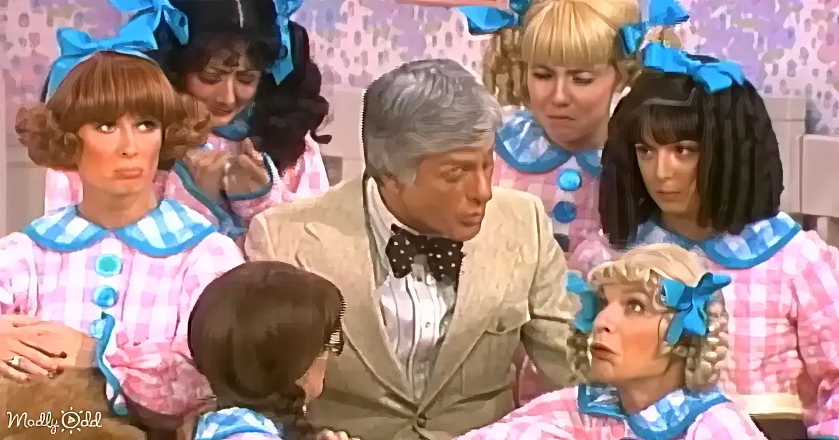 A man in a suit sits surrounded by women in pink and blue costumes, all with bows in their hair.