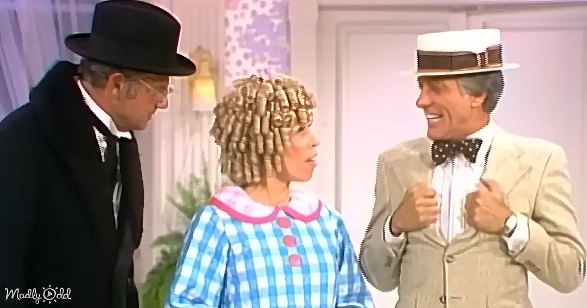 A woman in a blue checkered dress with a pink collar stands between a man in a black suit and a man in a beige suit with...
