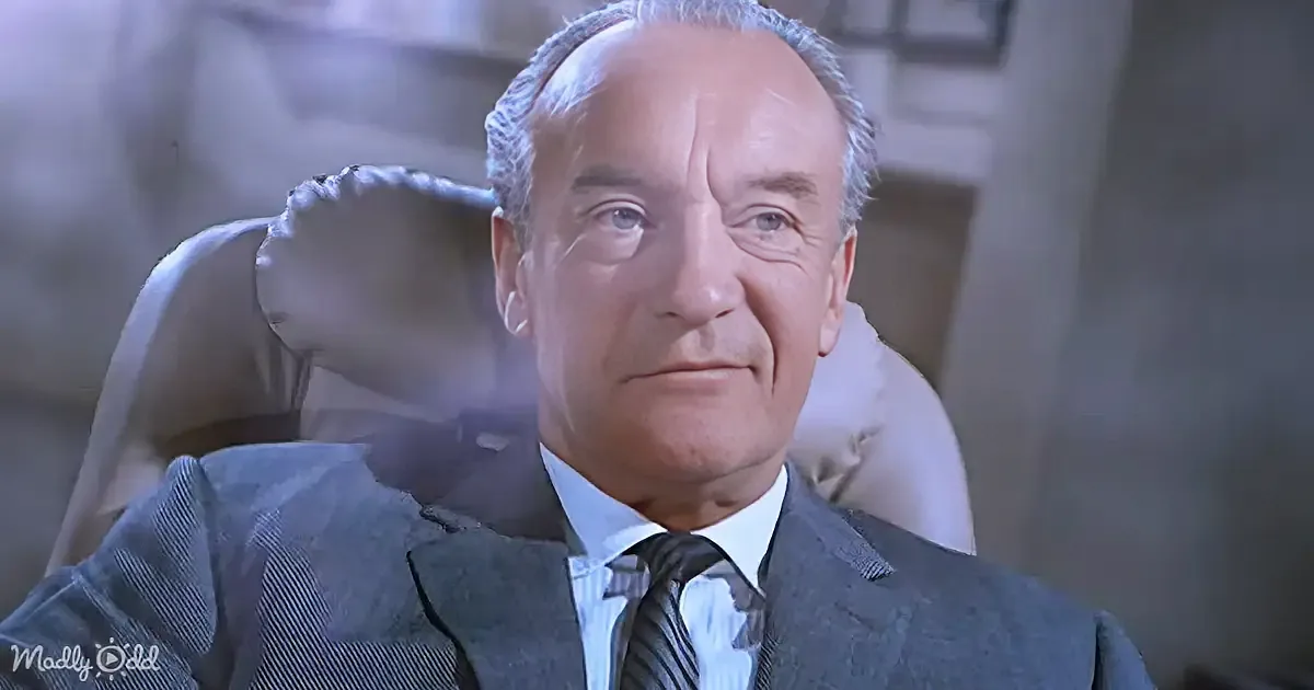 A man in a suit and tie sits in a chair, looking slightly to the side with a calm expression.