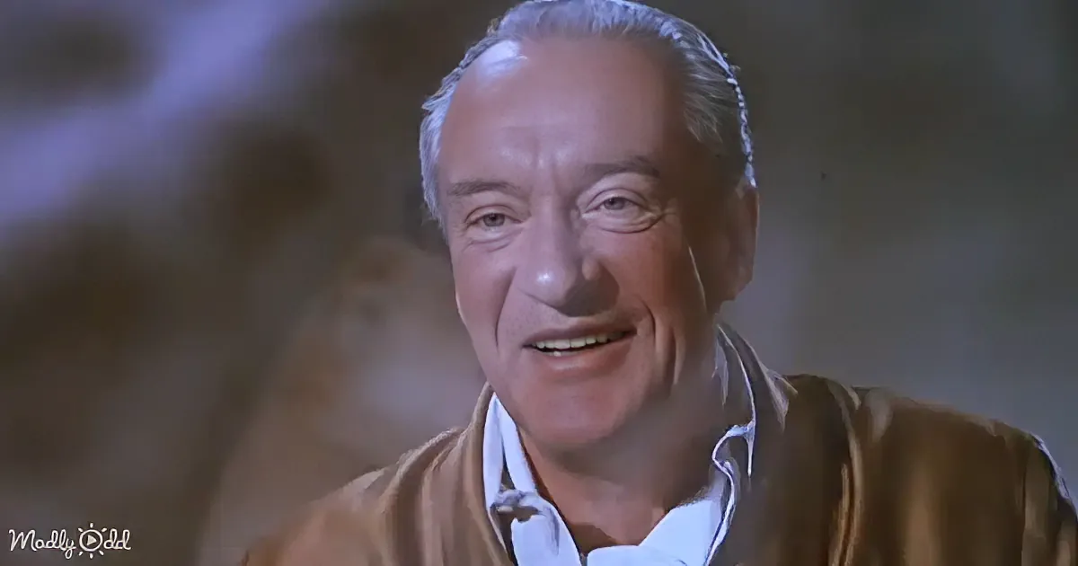 An older man with white hair, wearing a brown jacket and white shirt, smiles warmly.