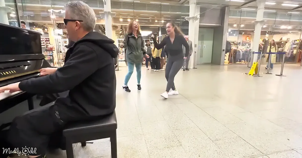 A man plays piano in a shopping mall as two women dance nearby.