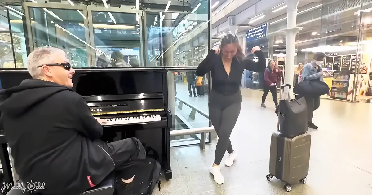 A man plays piano in a station while a woman dances nearby, with a woman sitting on a suitcase in the background.