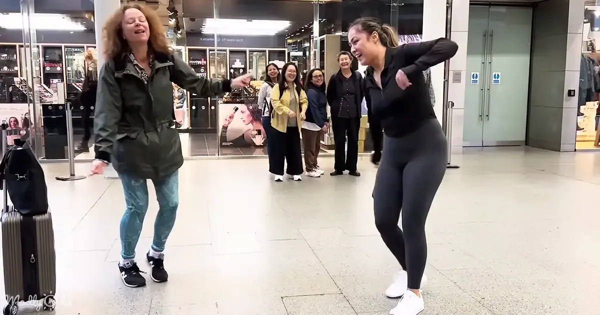 Two women dance joyfully in a shopping mall, surrounded by onlookers.