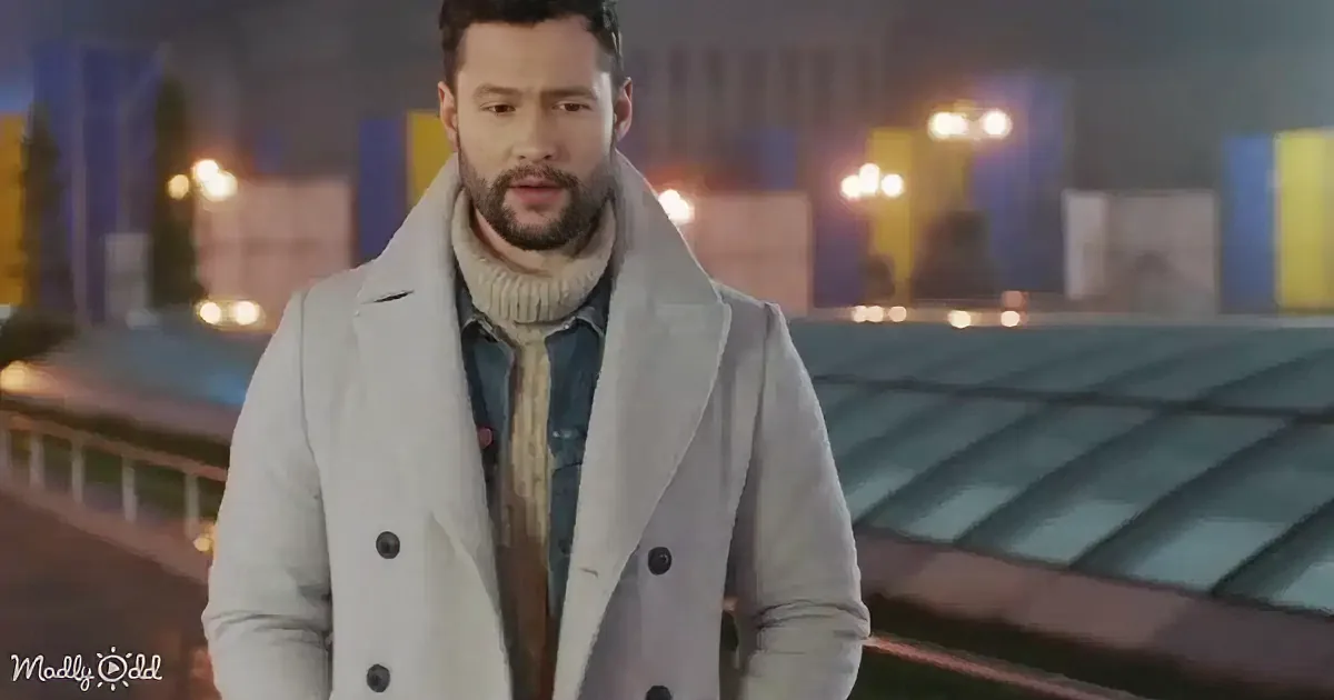 A man in a light gray coat and scarf stands on a bridge at night, with city lights and buildings in the background.