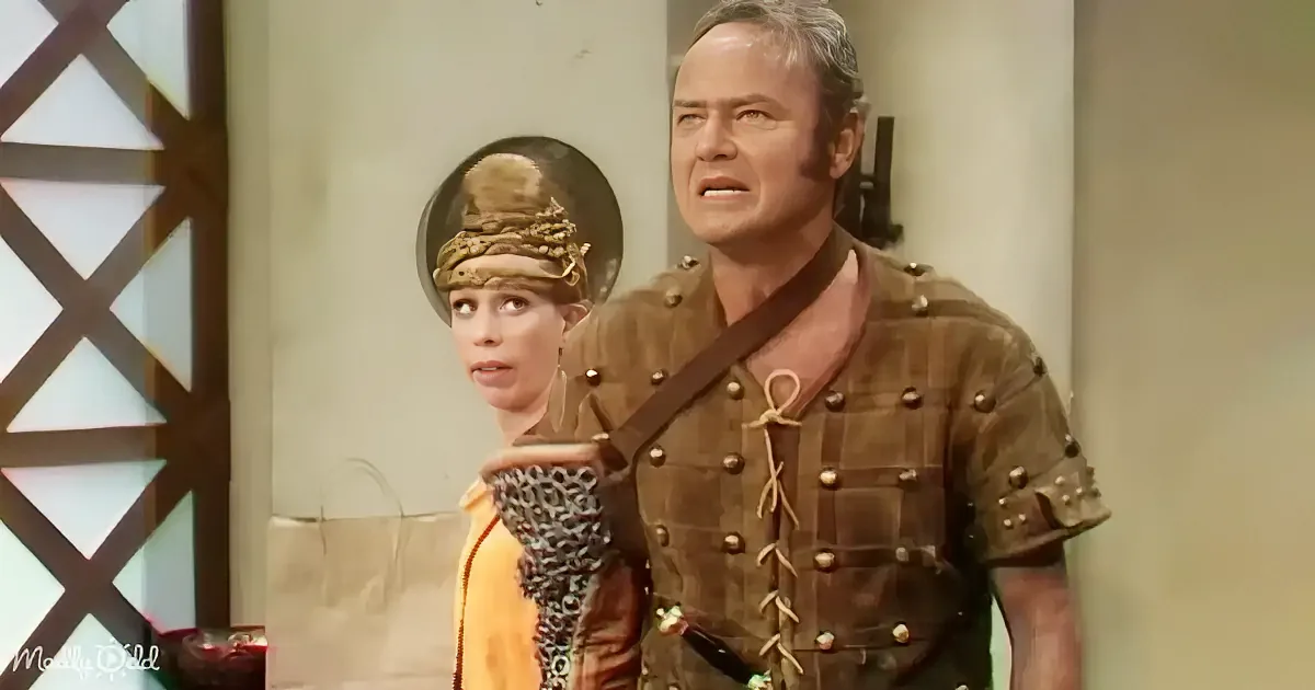 A man in a medieval-style tunic and armor stands next to a woman in a yellow dress and ornate headpiece.