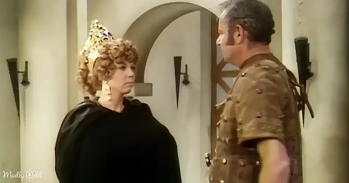A woman in a black outfit with a crown stands facing a man in a brown uniform with a sword, in a medieval setting.