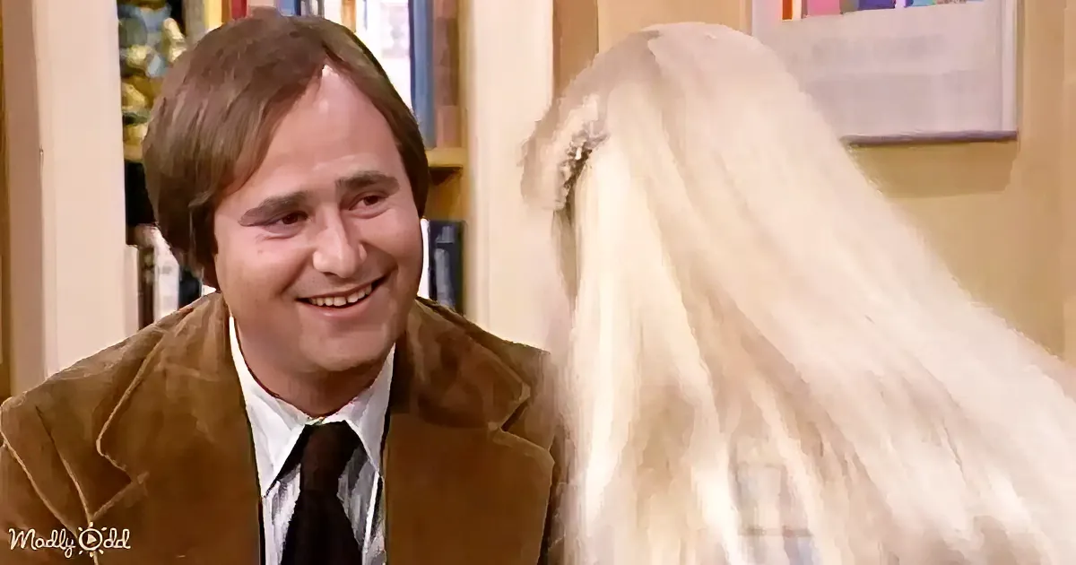 A man in a brown jacket and tie smiles at a person with long white hair.