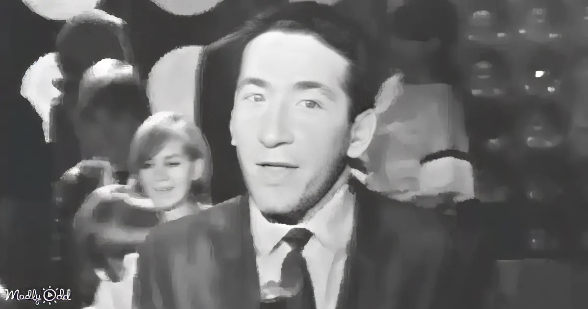 A man in a suit and tie is speaking, with blurred figures and lights in the background.