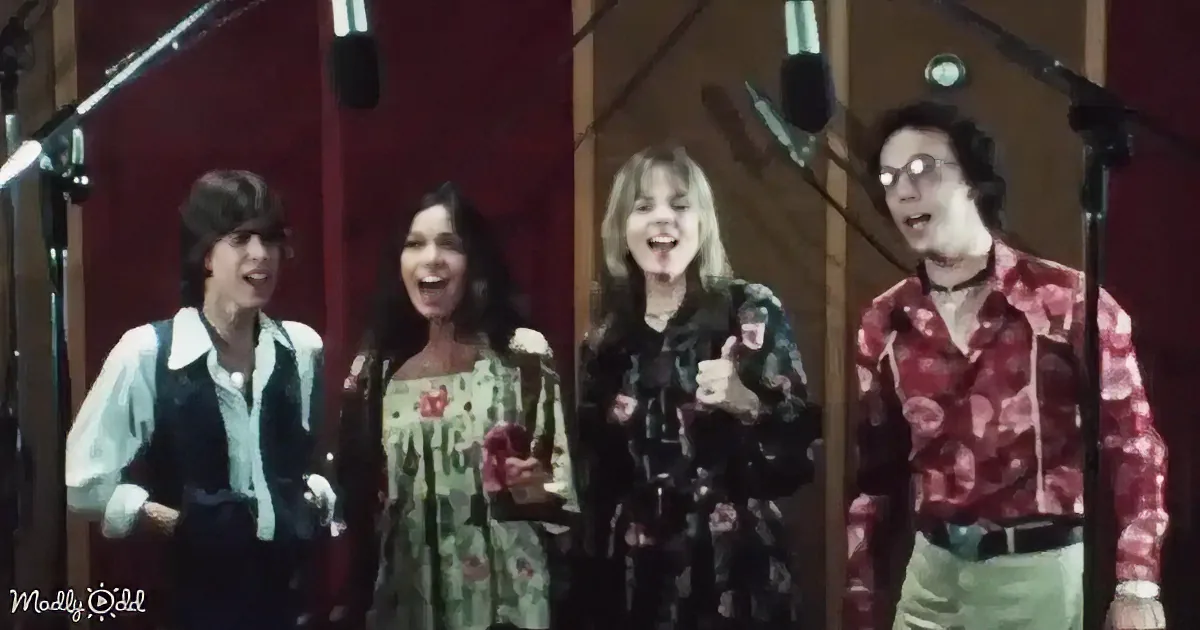 Four people in a recording studio, smiling and singing into microphones, wearing colorful 70s-style clothing.