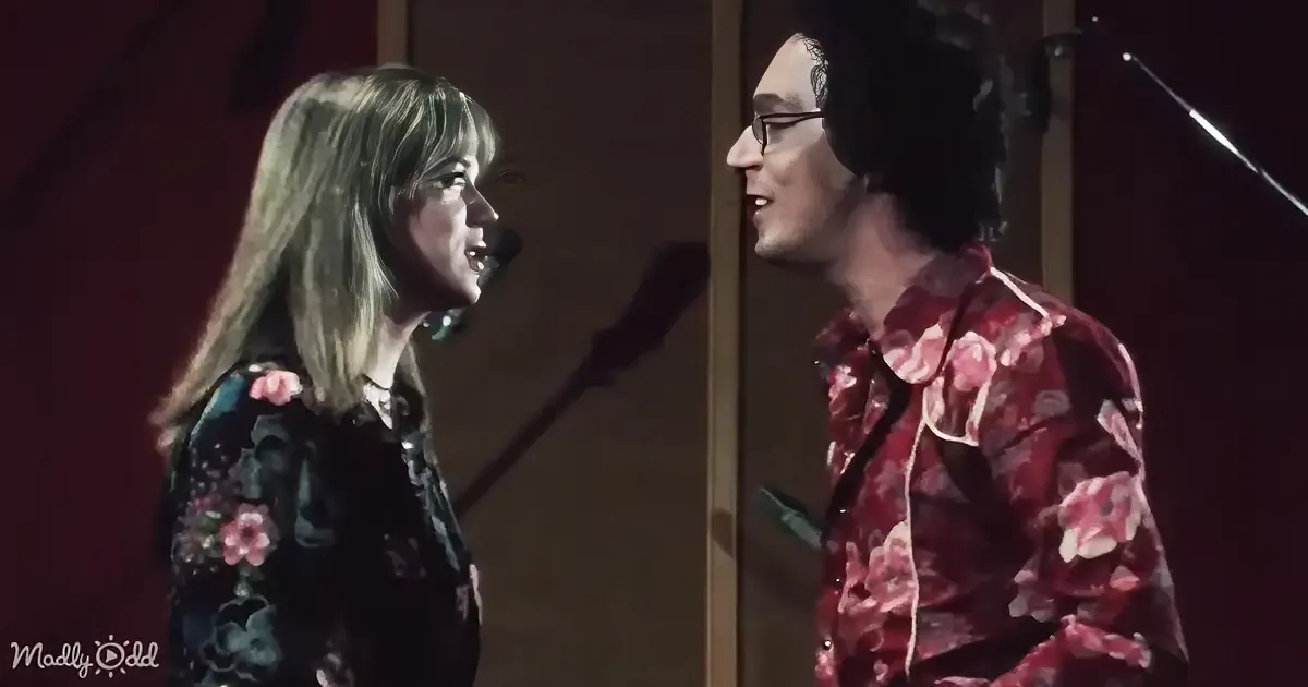 A woman in a floral dress sings into a microphone, facing a man in a similar floral shirt.